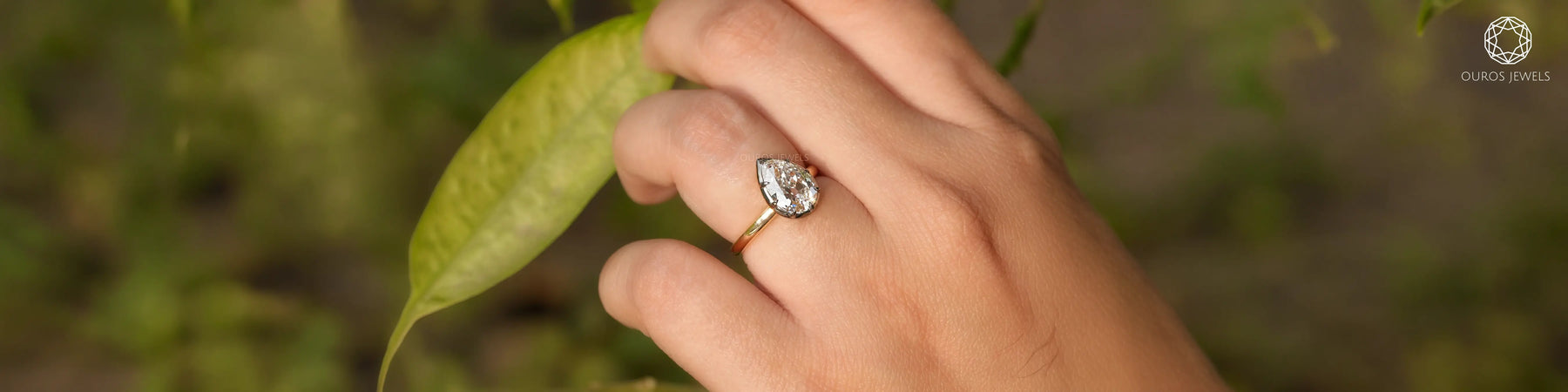 Pear shaped diamond engagement ring on petite hand with green leaf background by Ouros Jewels