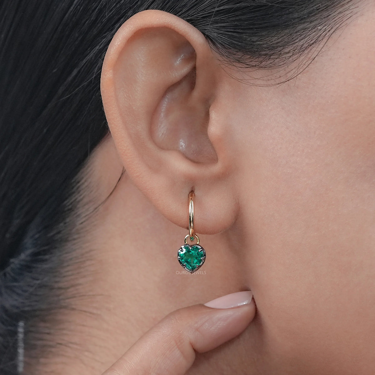 Heart shaped green emerald dangle huggie hoop earrings in yellow gold hinged setting.
